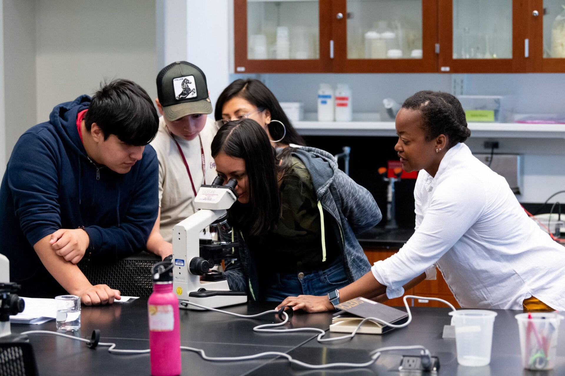 Three students, two men and one woman, look on as another female student looks through a microscope in a science lab while another woman looks on.