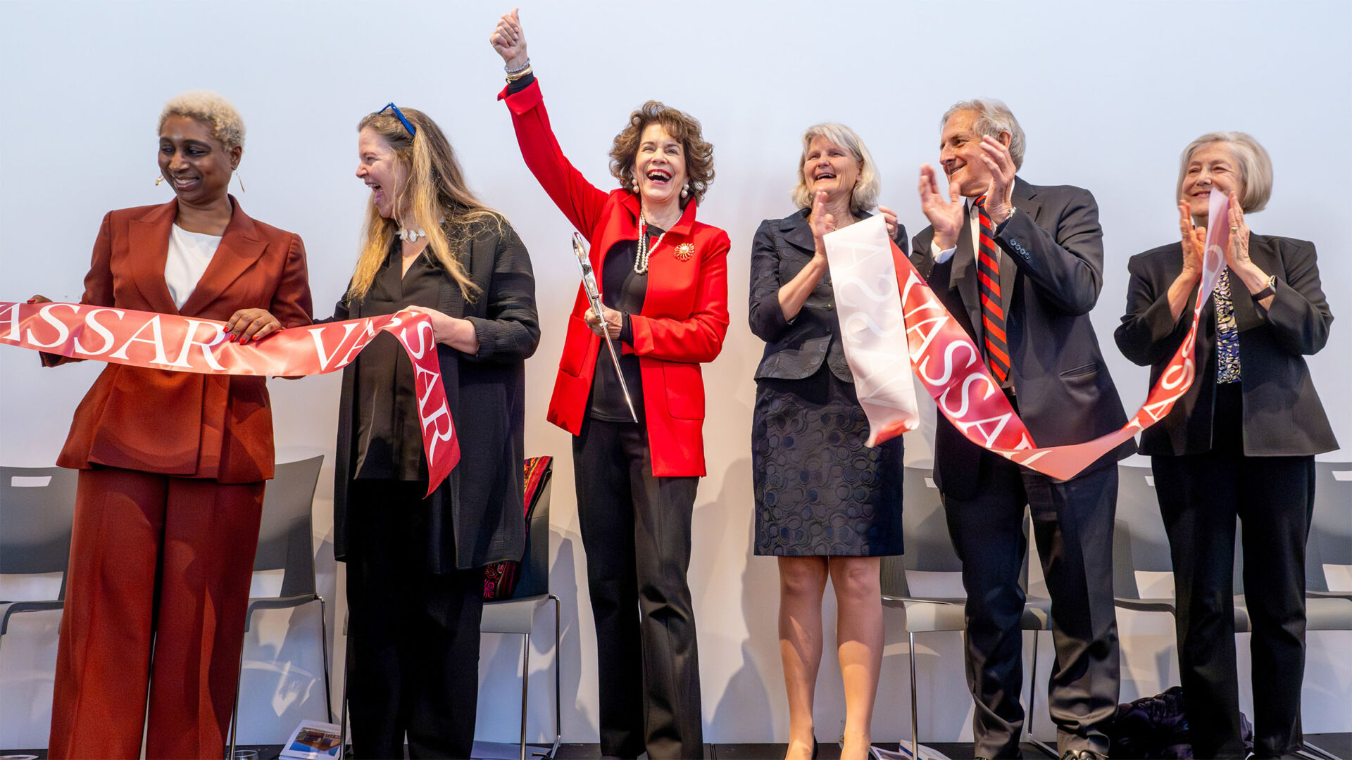 Six people celebrate a ribbon-cutting on stage. One person in a red jacket holds up scissors while the others cheer and hold a Vassar ribbon.